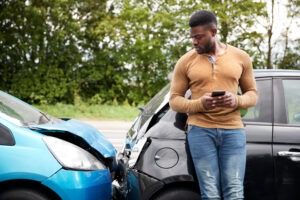 A man at a car accident scene uses his phone to research what happens if an unlicensed driver crashes your car.